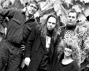 Four adults pose together outdoors in front of large leaves. Three stand in the back, one woman sits in front. All are casually dressed and smiling or relaxed, in a black and white photo.