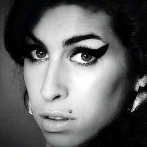 A close-up black and white photo of a woman with thick, winged eyeliner, arched eyebrows, a beauty mark above her lip, hoop earrings, and a serious expression, looking directly at the camera.