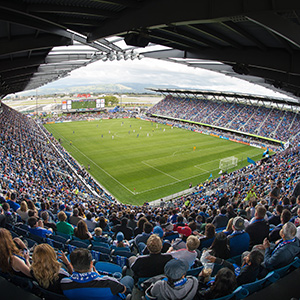 A wide-angle view of a packed soccer stadium, with fans filling the stands and a match underway on the green field under a partially cloudy sky. The photo is taken from high in the stands behind the goal.