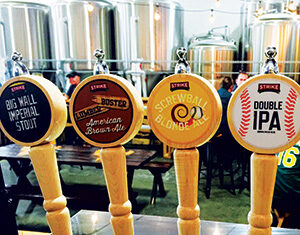 Four wooden beer taps labeled Big Wall Imperial Stout, Lumber Buster American Brown Ale, Screwball Blonde Ale, and Double IPA, with stainless steel brewing tanks in the background.