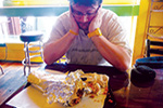 A person sits at a table, staring down at a very large burrito partially wrapped in foil, looking thoughtful or overwhelmed. Bright, colorful café surroundings are visible in the background.