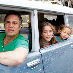 A man and two young girls sit in an old blue car, looking out of the open window. The man is in a green shirt, while the girls sit beside him in the back seat, gazing attentively at something outside.
