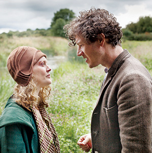 A woman in a brown hat and coat faces a man in a brown jacket outdoors, standing close together in a grassy field with trees in the background under a cloudy sky.