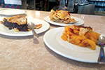 Three white plates each hold a slice of pie on a countertop; one looks to be chocolate, another appears to be apple, and the third is possibly a fruit pie. Spoons rest on two of the plates.