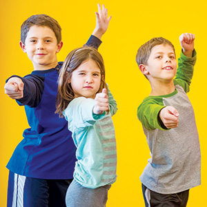 Three children stand in dynamic, playful poses pointing forward, smiling confidently against a bright yellow background.