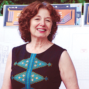 A woman with curly brown hair smiles at the camera. She is wearing a sleeveless black top with blue and gold patterns. Behind her are posters with architectural drawings and greenery at the top.
