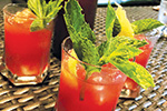 Three glasses filled with a bright red drink, garnished with fresh green mint leaves and lemon peel, are placed on a woven tabletop. The beverages appear to be refreshing cocktails or mocktails.