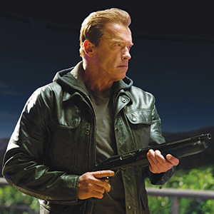 A man in a black leather jacket holds a shotgun and looks to the side with a serious expression, standing outdoors at night.