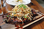 Grilled steak topped with caramelized onions and herbs, served on a wooden board with a glossy sauce; a plate of rice is in the background.