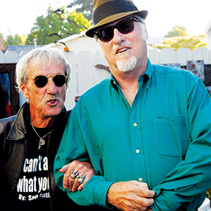 Two older men stand together outdoors. Both wear sunglasses; one has a black jacket and t-shirt, the other wears a teal button-up shirt and fedora. They appear friendly and relaxed, with one linking arms with the other.