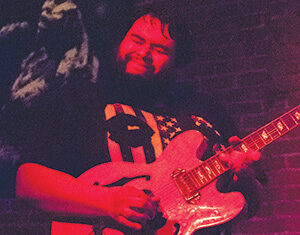 A bearded man plays an electric guitar passionately on stage under red lighting, wearing a black T-shirt with an American flag design. A brick wall is visible in the background.