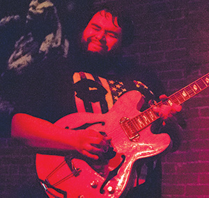 A bearded man plays an electric guitar passionately on stage under red lighting, wearing a black T-shirt with an American flag design. A brick wall is visible in the background.
