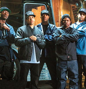 Five men stand in front of a bus, wearing dark jackets and N.W.A hats, with serious expressions and arms crossed, posing together in an urban nighttime setting.