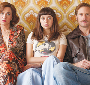 Three people sit on a couch against a retro patterned wallpaper. The woman on the left wears a floral outfit, the young woman in the center wears a t-shirt and jeans, and the man on the right wears a brown jacket and jeans.