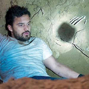 A man with a beard and messy hair, wearing a dirty white shirt, sits against a cracked, dirt wall. A skeletal hand imprint and a shadow are visible on the wall next to him.