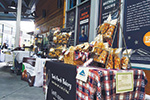 Outdoor market stall with tables covered in plaid cloths displaying packaged baked goods and treats. A person stands behind the display, and signs with information are visible on the wall in the background.