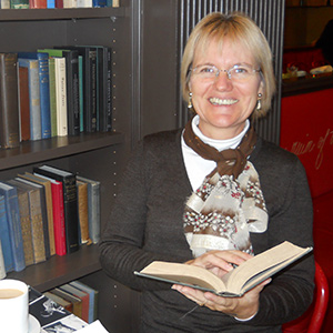 A smiling woman with glasses sits by a bookshelf, holding an open book. She is wearing a brown sweater and a patterned scarf. A cup of coffee and papers are on the table in front of her.
