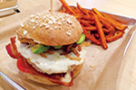 A burger with bacon, a fried egg, avocado, and onions on a seeded bun sits on a tray next to a serving of sweet potato fries.