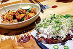 A bowl of fried chicken bites with green garnish next to a slice of bread topped with shredded cheese, sprouts, and sliced green onions on a wooden table.