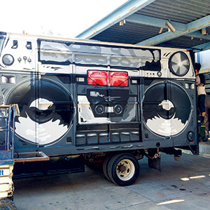 A truck painted to look like a large vintage boombox, complete with speakers, dials, and cassette deck details, parked under an open metal roof structure.