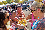Four women stand in a sunny outdoor market, sharing food from a tray and smiling. White tents and a crowd are visible in the background, suggesting a lively festival atmosphere.