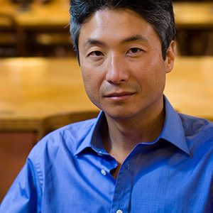 A man with short, dark hair wearing a blue collared shirt sits indoors, looking at the camera with a neutral expression. The background is softly blurred, showing wooden tables and chairs.