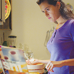 A woman in a purple shirt stands in a kitchen looking at a cookbook while preparing food, with her hands near a mixing bowl on the counter.