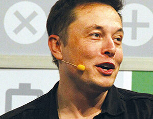 A man with light skin and short brown hair wears a black shirt and a headset microphone, smiling while looking to the side. The background features large symbols, including an X and a battery icon.