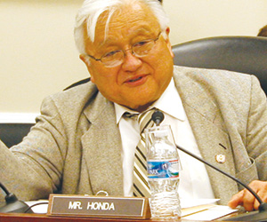 An older man with white hair and glasses sits at a desk wearing a gray suit and striped tie. A nameplate reading Mr. Honda and a water bottle are in front of him. He appears to be speaking or gesturing.