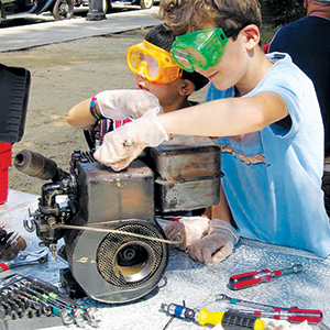 Two children wearing safety goggles and gloves work together to repair or examine a small engine on a table covered with tools outdoors.