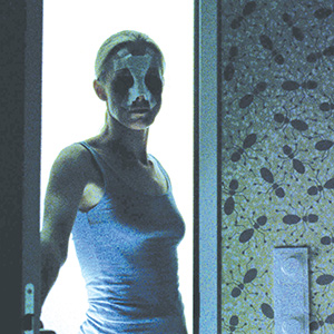 A person wearing a sleeveless top and a facial bandage stands in a doorway, backlit by bright light, with a patterned wall and door handle visible on the right.