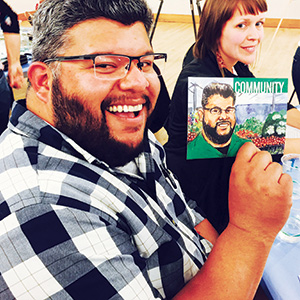 A smiling man in glasses and a plaid shirt holds up a drawing of himself labeled Community while sitting at a table. A woman with red hair sits beside him, also smiling.