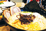 A close-up of a bowl of ramen with sliced pork, seaweed, noodles, mushrooms, and green onions in a rich broth, served on a table.