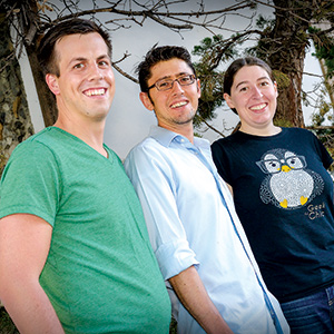 Three people stand outdoors smiling at the camera. The person on the left wears a green shirt, the middle person wears glasses and a light blue shirt, and the person on the right wears a black shirt with an owl graphic.
