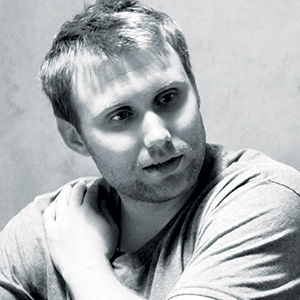 A man with short, light hair and stubble looks to the side, resting one hand on his shoulder. He is wearing a t-shirt, and the image is in black and white with soft lighting.