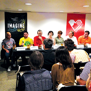 A panel of seven people sits at a long table facing an audience in a conference room. Two banners, one reading IMAGINE, hang behind them. A man on the left speaks into a microphone.