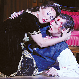 A woman in a black dress cradles a wounded man with blood on his face, sitting on the floor by a red chair, both with dramatic expressions, suggesting a tense or emotional scene.