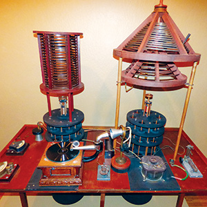 A vintage electrical apparatus with coils, switches, and gears on a wooden table, featuring two large circular components and a sloped conical structure, evoking early 20th-century scientific equipment.