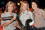 Three women in matching khaki shirts smile at the camera while holding a large snake together at what appears to be an indoor event or exhibit.