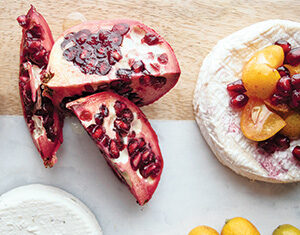 Sliced pomegranate, whole kumquats, and two rounds of cheese—one topped with kumquats and pomegranate seeds—are arranged on a light-colored surface.