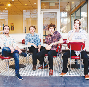 Four young men sit on red chairs around white tables in a retro-style diner with checkered tile floor and large windows. They all wear patterned shirts and look relaxed, posing for the camera.
