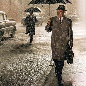 A man in a trench coat and hat walks on a rainy, city street at night holding an umbrella. Another person with an umbrella walks in the background, and vintage cars are parked along the wet sidewalk.