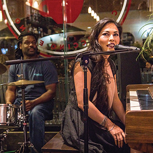 A woman sings and plays piano at a microphone, while a man plays drums behind her in a cozy, warmly lit venue with a large window in the background.