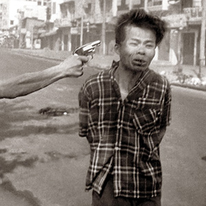 A black and white photo shows a man in a plaid shirt with his hands tied behind his back as another person points a handgun at his head on a city street, with blurred buildings visible in the background.