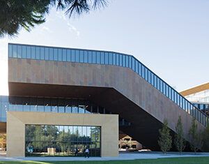 Modern building with large glass windows and an angular, cantilevered upper floor, surrounded by green lawn and trees under a clear sky.