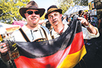 Two men wearing traditional Bavarian attire hold a large German flag at an outdoor festival. One man is holding a beer stein, and both are smiling. There are people and tents in the background.