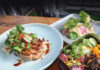 A plated grilled meat patty topped with fresh herbs and sauce sits next to a bento box containing sliced raw fish, vegetables, and pickled items on a wooden table.