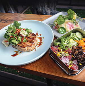 A plated grilled meat patty topped with fresh herbs and sauce sits next to a bento box containing sliced raw fish, vegetables, and pickled items on a wooden table.