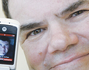 A man smiles closely at the camera while holding an open flip phone, which displays his own face on its screen.