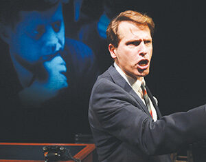 A man in a suit passionately speaks on stage with a serious expression. Behind him, a large projected image of another man, also in a suit, is shown resting his chin on his hand in a thoughtful pose.